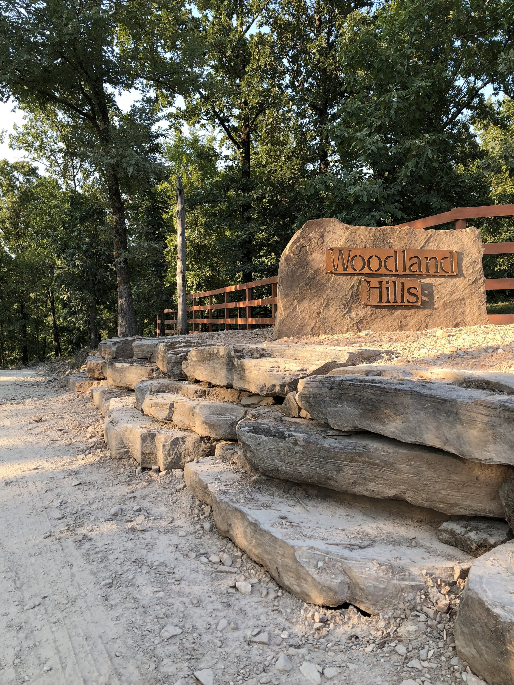 A large rock with a sign that reads 'Woodland Hills' engraved into it. The rock is part of a stone retaining wall, and behind it there is a wooden fence. Trees with green leaves are visible in the background.