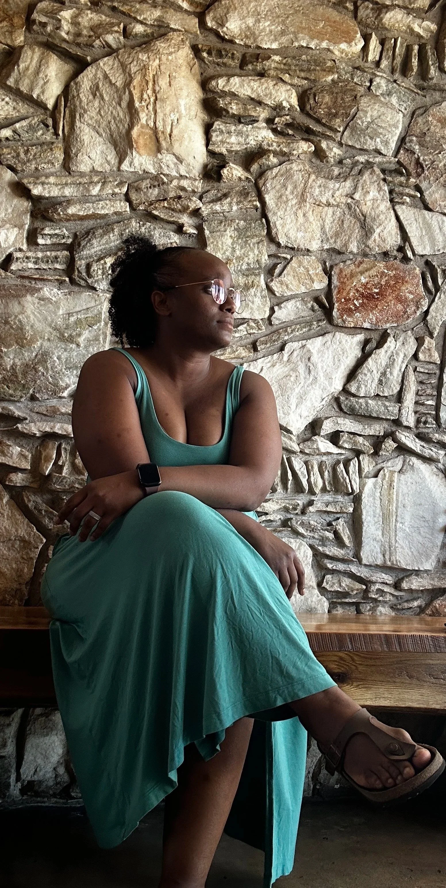 A woman with glasses and a black smartwatch sitting on a wooden bench against a stone wall, wearing a teal dress and sandals.