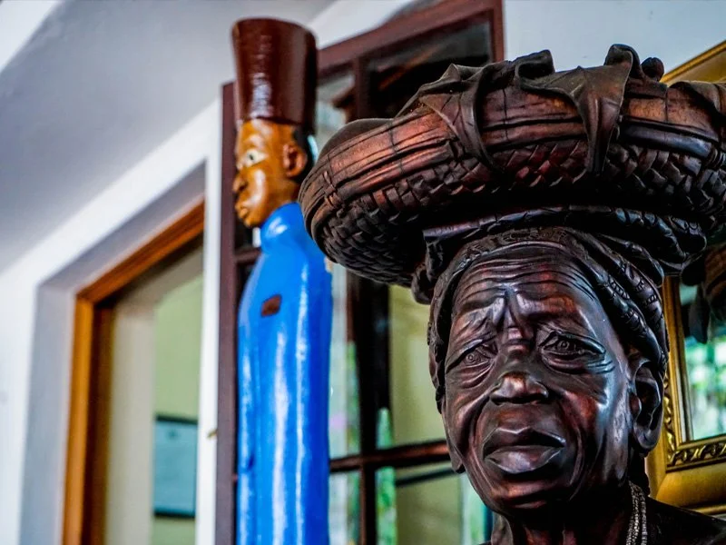 Detailed close-up of a dark wooden sculpture of an elderly woman with a head wrap, displaying expressive facial features, with a blurred sculpture of a woman in a blue dress and a tall hat in the background.