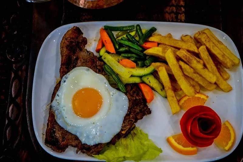 A plate of food with a fried egg on top of a cooked steak, served with French fries, mixed vegetables, orange slices, and a decorative tomato carved into a rose.