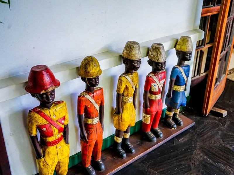 A row of five vintage wooden figurines dressed in colorful military uniforms and hats, standing on a wooden base against a wall.