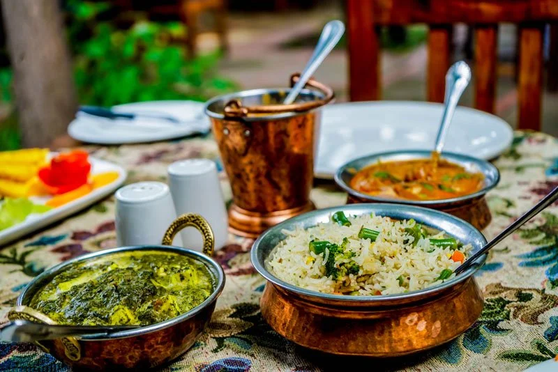 Indian meal served on a table with rice, curry, and vegetable dishes in copper bowls, set on a floral tablecloth outdoors.