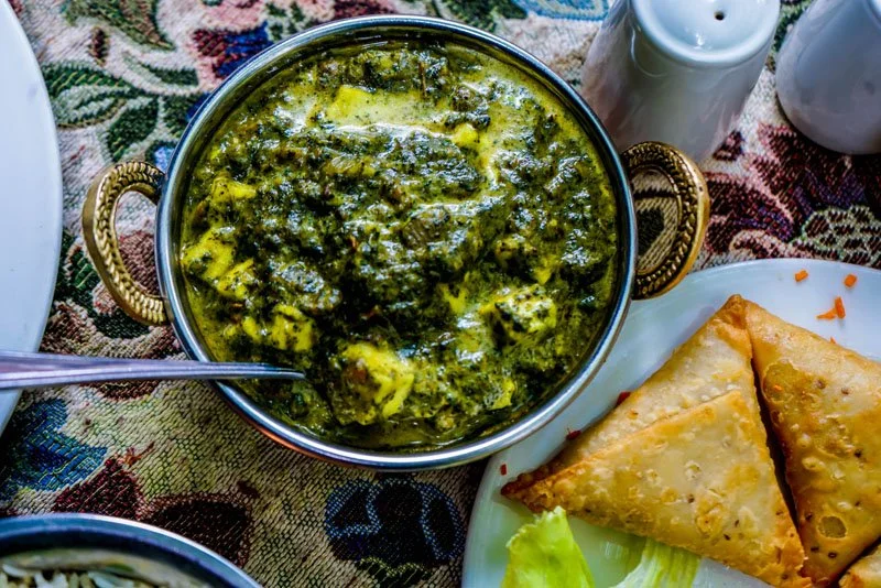 A metal bowl of Indian spinach and cheese dish, served with crispy fried samosas on a plate with a lettuce leaf, salt, and pepper shakers in the background, on a patterned tablecloth.