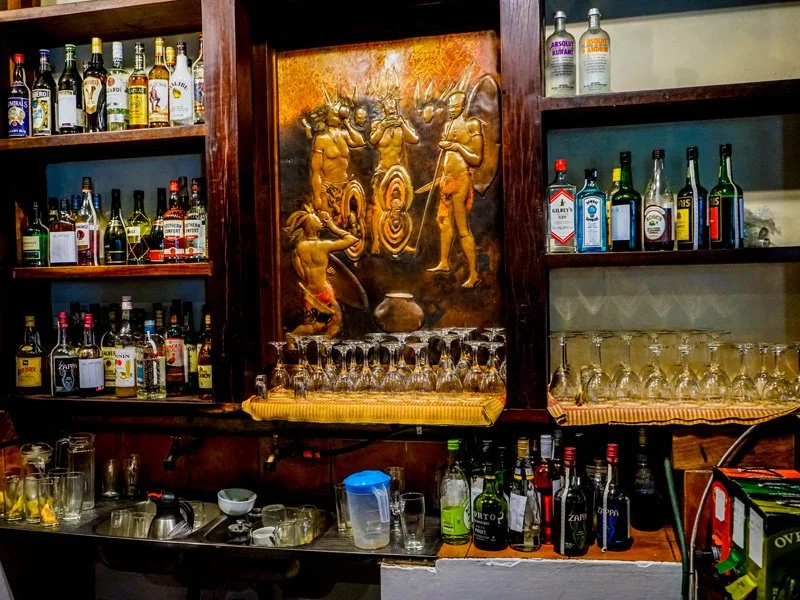 A bar with shelves of various liquor bottles, glassware, and a decorative wooden carving depicting traditional figures.