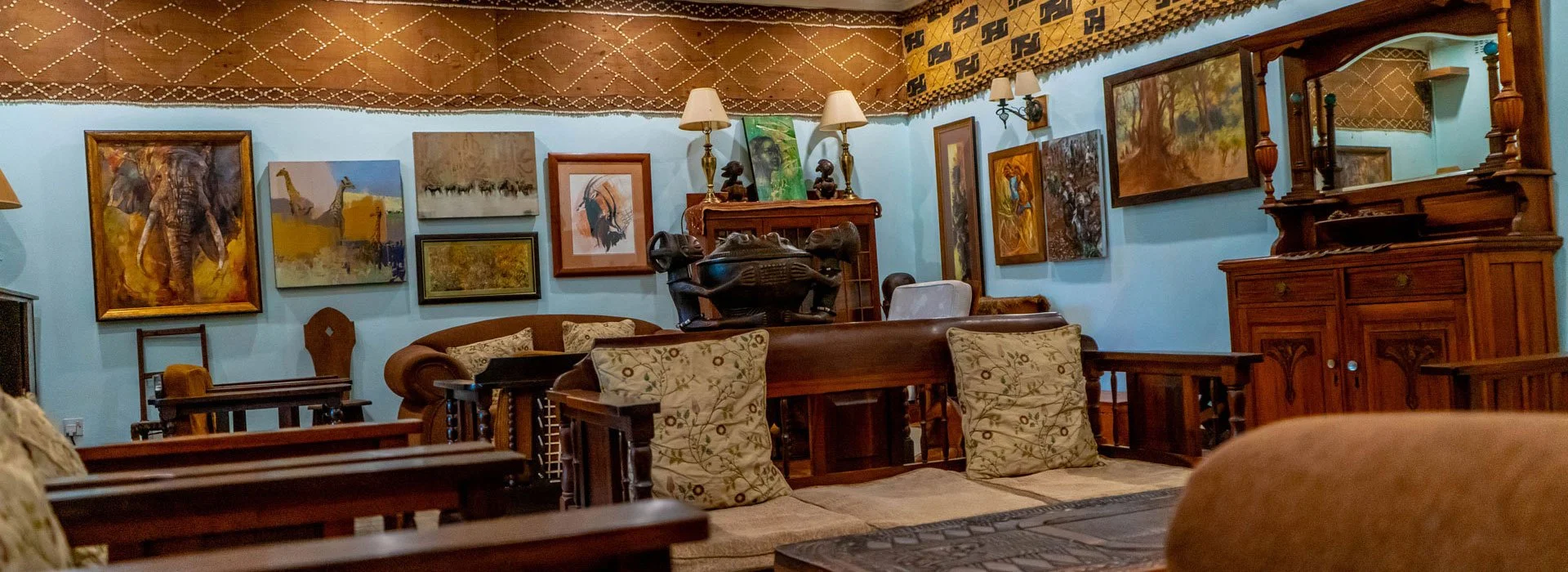 Living room with light blue walls decorated with numerous paintings and framed artwork, wooden furniture including a cabinet and a sofa with beige and patterned cushions, lamps on top of a wooden shelf, and various traditional artifacts and sculpture