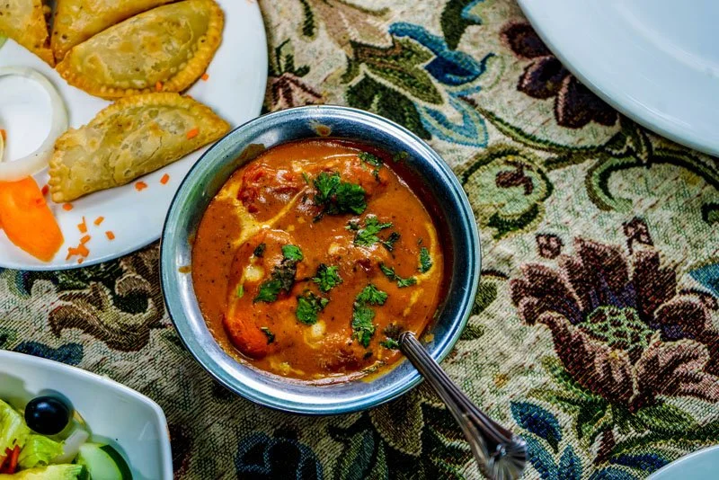 Indian curry with cilantro garnishing in a metal bowl, served with vegetable samosas and salad on a decorative tablecloth.