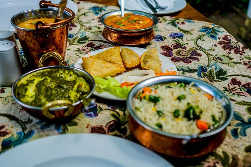 Indian meal with various dishes including a copper bowl of dal, a copper bowl of vegetable curry, a plate of samosas, a bowl of spinach, and a bowl of vegetable rice on a floral tablecloth.
