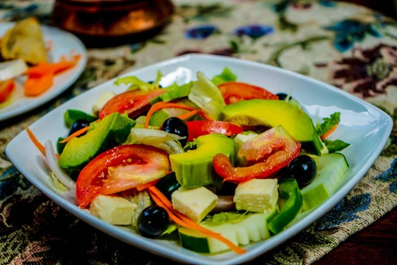 Fresh vegetable salad with tomatoes, avocado, black olives, cucumber, carrots, and cheese in a white bowl