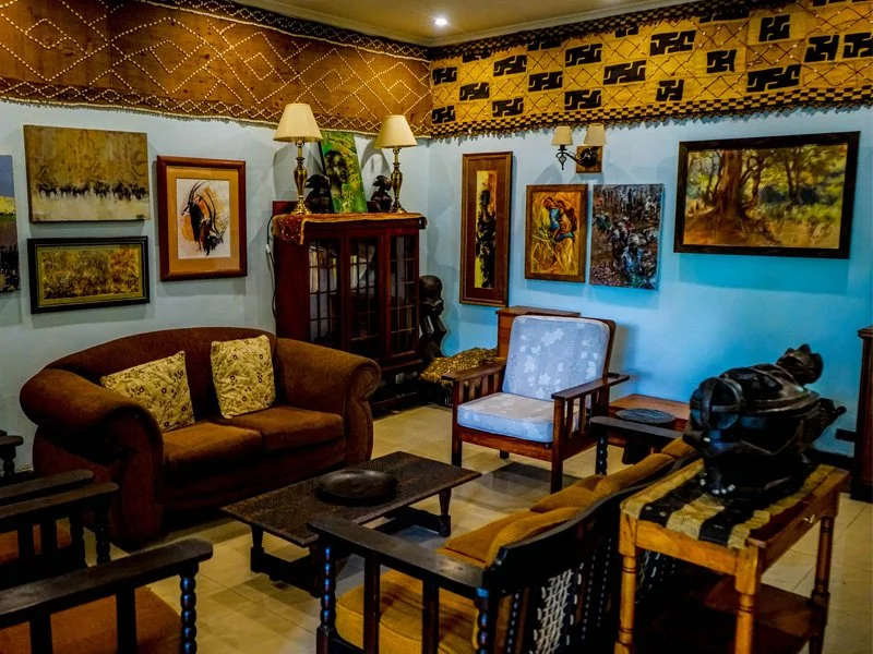 Living room with brown sofa, wooden armchair, blue upholstered armchair, paintings on the walls, a wooden display cabinet, and antique items.