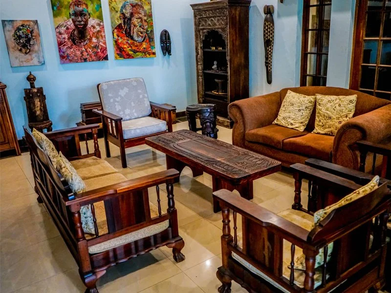 Living room with wooden furniture including armchairs, a coffee table, and a sofa with patterned pillows, decorated with colorful paintings on the blue wall and wooden shelves.