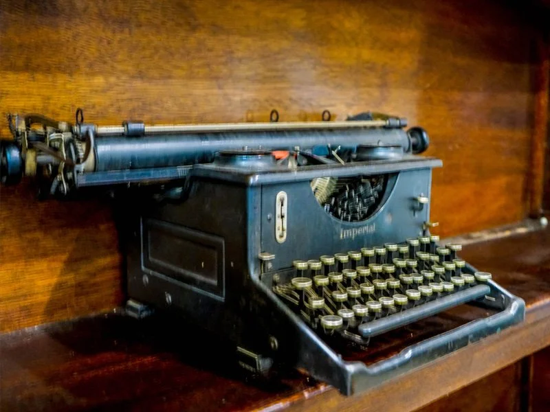 A vintage black Imperial typewriter with round keys placed on a wooden surface against a wooden wall.