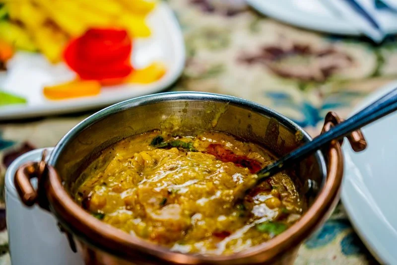 A pot of yellow, thick lentil curry with herbs on a table, with a plate of sliced vegetables in the background.