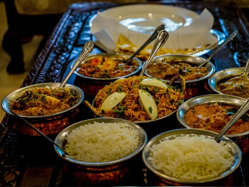 Indian feast with bowls of rice, curry dishes, and a plate of vegetable pulao, on a dark table.
