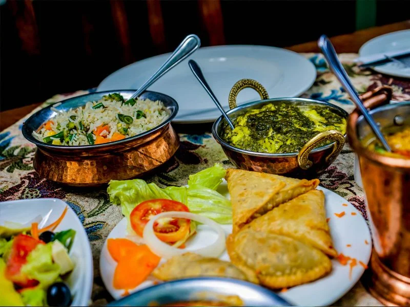 A table with Indian food, including a salad with tomatoes, onions, and lettuce, a dish of rice, a bowl of spinach or green curry, and samosas, with utensils and empty plates.