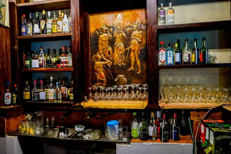 Bar shelf with various liquor bottles, glasses, and a decorative bronze relief in the center depicting four traditional dancers with drums, with no people present.