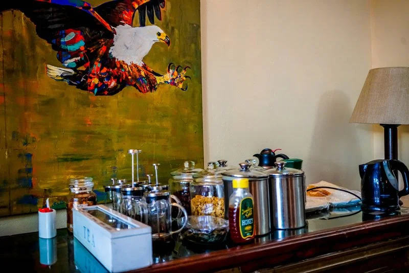 A breakfast or snack setup on a dark wooden table with jars of cereal, a bottle of honey, a teapot, a kettle, and a lamp. A colorful painting of a flying eagle is on the wall behind.