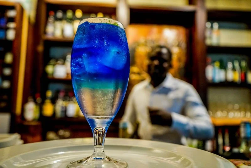 Colorful layered cocktail in a tall glass with a blue top layer and a clear bottom layer, served on a white tray, with a blurry bartender and bar shelves in the background.