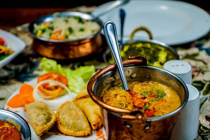 A variety of Indian dishes on a table, including a pot of yellow lentil curry, samosas, and vegetable dishes.