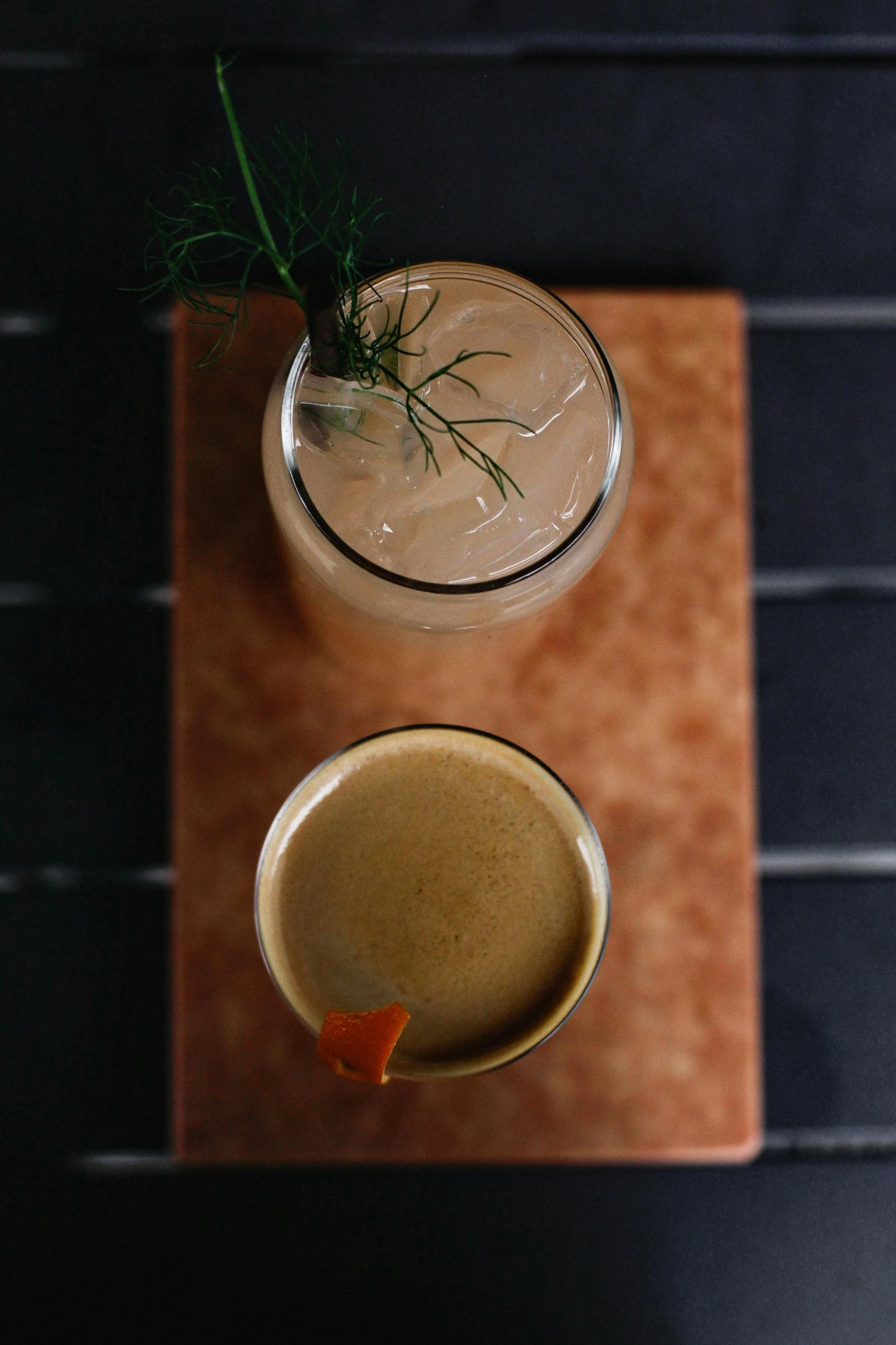 A top-down view of two beverages on a wooden tray against a dark background. One drink is garnished with a small leafy sprig and ice, and the other has a small orange peel on the rim.