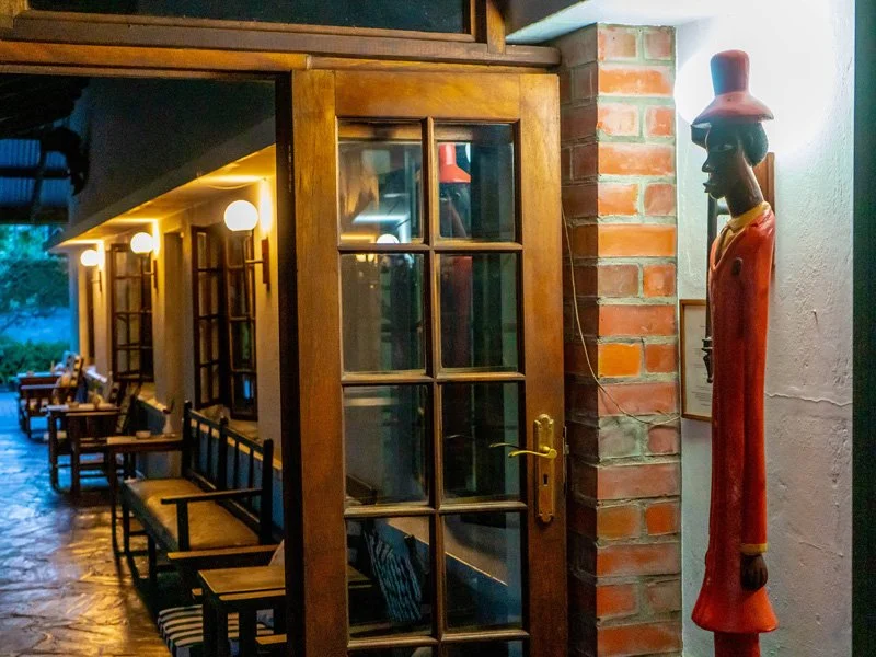 Interior view of a cozy restaurant or café with wooden furniture, a brick wall, and warm ambient lighting. A carved wooden statue of a figure in a red coat and hat is mounted on the wall near a glass-paneled wooden door.