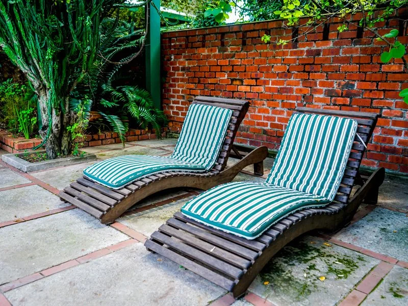 Two wooden lounge chairs with green and white striped cushions on a patio with a brick wall and green plants in the background.