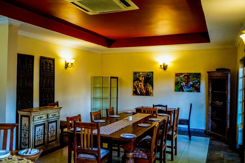 A dining room with a wooden table set for six, decorated with a table runner and place settings. The room has yellow walls, two framed paintings, and is lit by wall sconces. There are also two wooden cabinets and a window with natural light.