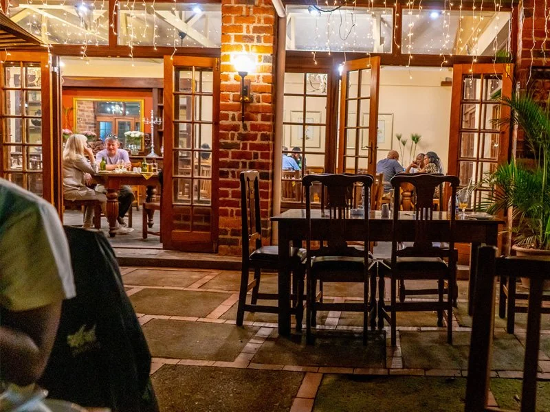 Outdoor patio of a restaurant with wooden tables and chairs, open glass-paneled doors revealing indoor dining, people eating and socializing, string lights, brick walls, and potted plants.