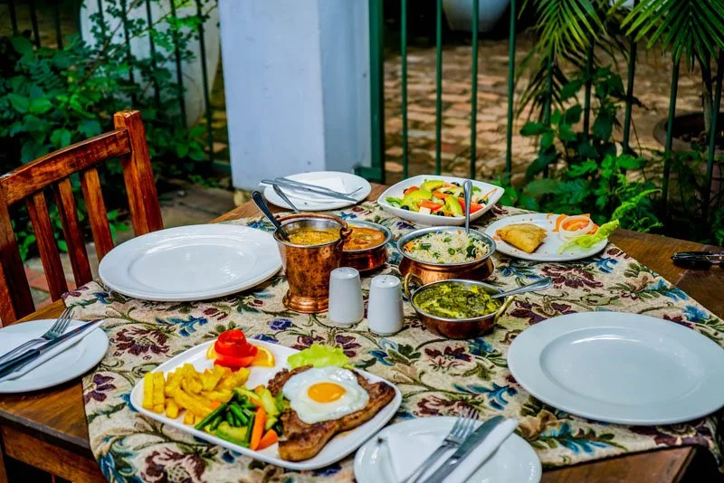 A table set for a meal with two empty plates, a fried egg on a meat cutlet, assorted vegetables, and various dishes, with utensils and salt and pepper shakers, outdoors with greenery and a fence in the background.