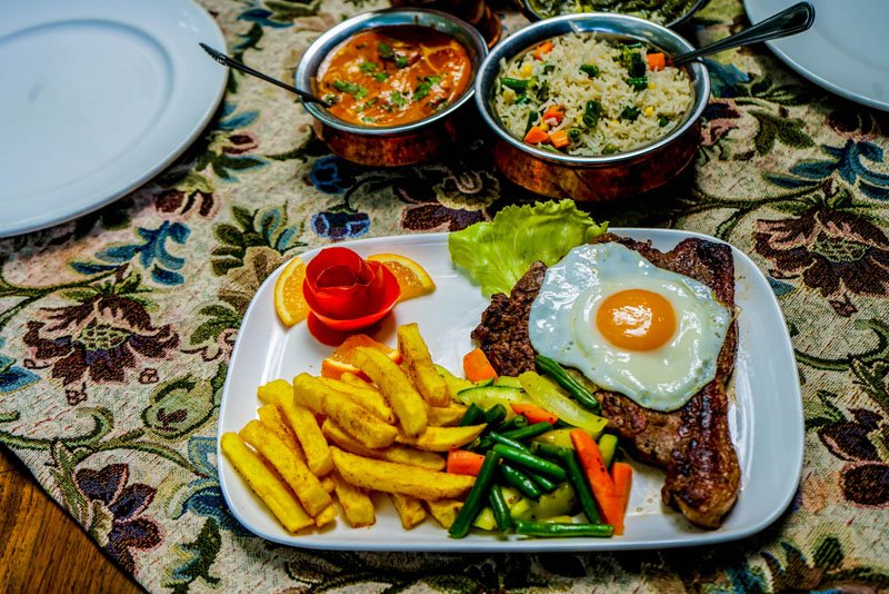A plate with fried plantains, a grilled steak topped with a sunny-side-up egg, mixed vegetables, and a decorative tomato rose. In the background, there are bowls of soup and rice. The table has a patterned tablecloth.