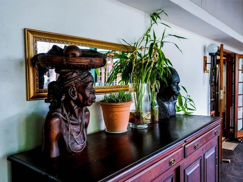 Wooden sideboard with framed mirror above, decorated with two bust sculptures, a potted plant, and a tall glass vase with greenery, in a home interior.