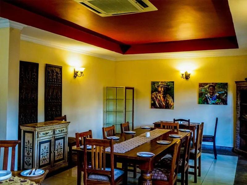 Dining room with a long wooden table set with plates, napkins, and a table runner, surrounded by wooden chairs. The room has yellow walls, decorative wood panels, and two paintings on the wall. There are wall sconces providing light, and a glass disp