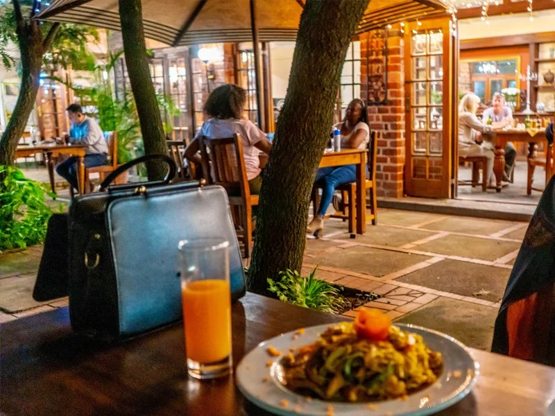 Outdoor restaurant patio with people dining, a table with food, drink, and a black handbag, trees, and warm lighting