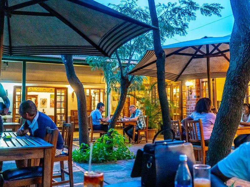 Patio dining area with people seated at wooden tables under umbrellas, surrounded by trees and plants, in front of a building with large windows and warm lighting.