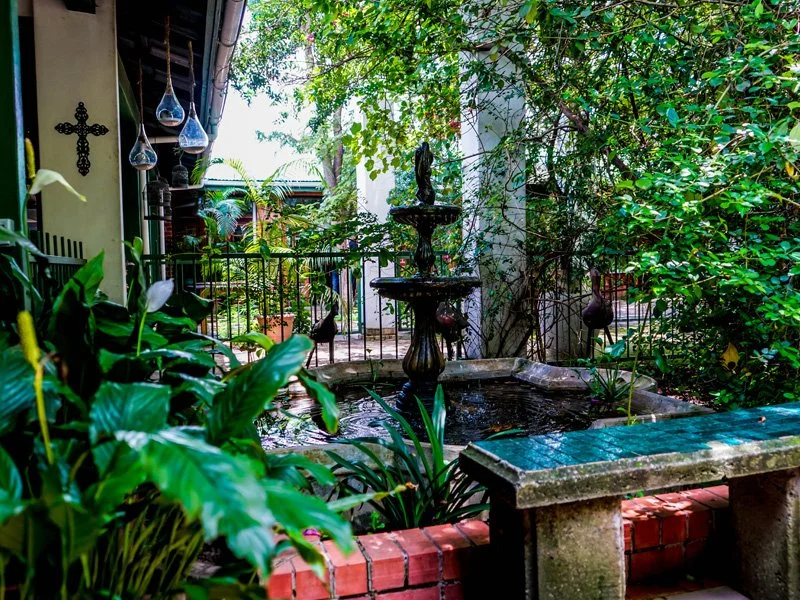A cozy garden patio featuring a small tiered fountain at its center, surrounded by lush green plants, trees, and decorative hanging glass ornaments. A brick ledge frames the fountain area.