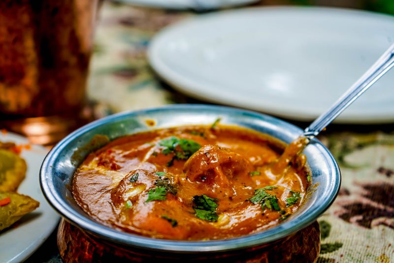 A bowl of Indian butter chicken curry garnished with cilantro, with a spoon, on a table with plates and a drink in the background.