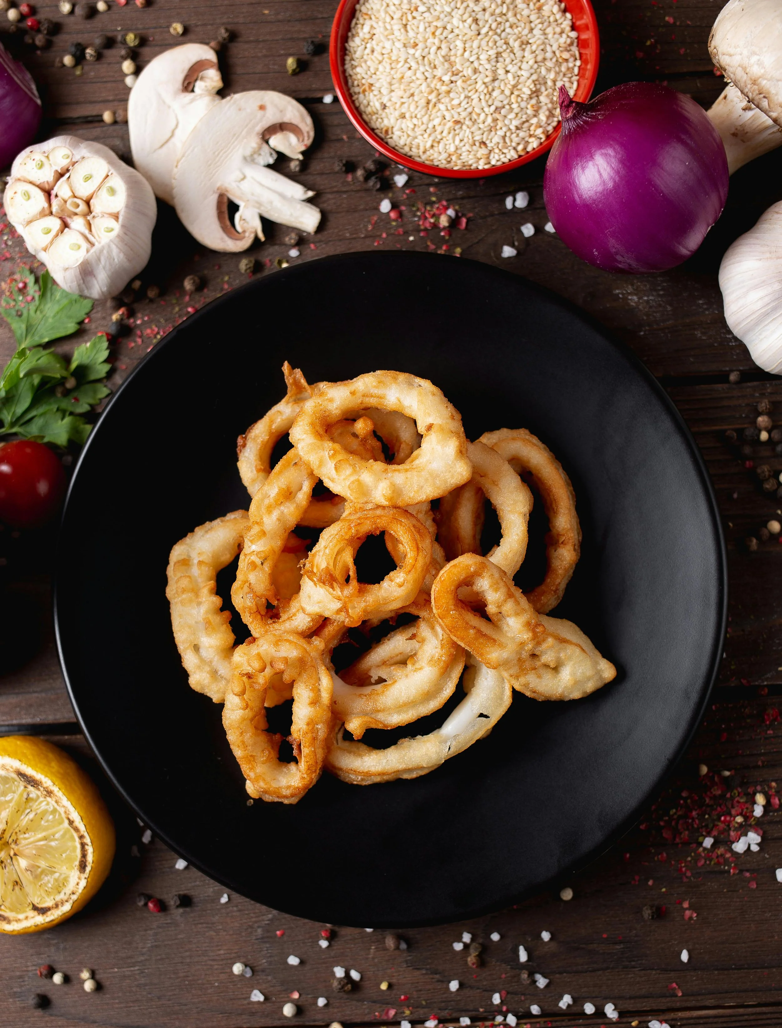 Fried calamari rings on black plate with garlic, lemon, red onion, mushrooms, garlic cloves, parsley, rice, and mixed peppercorns surrounding it.