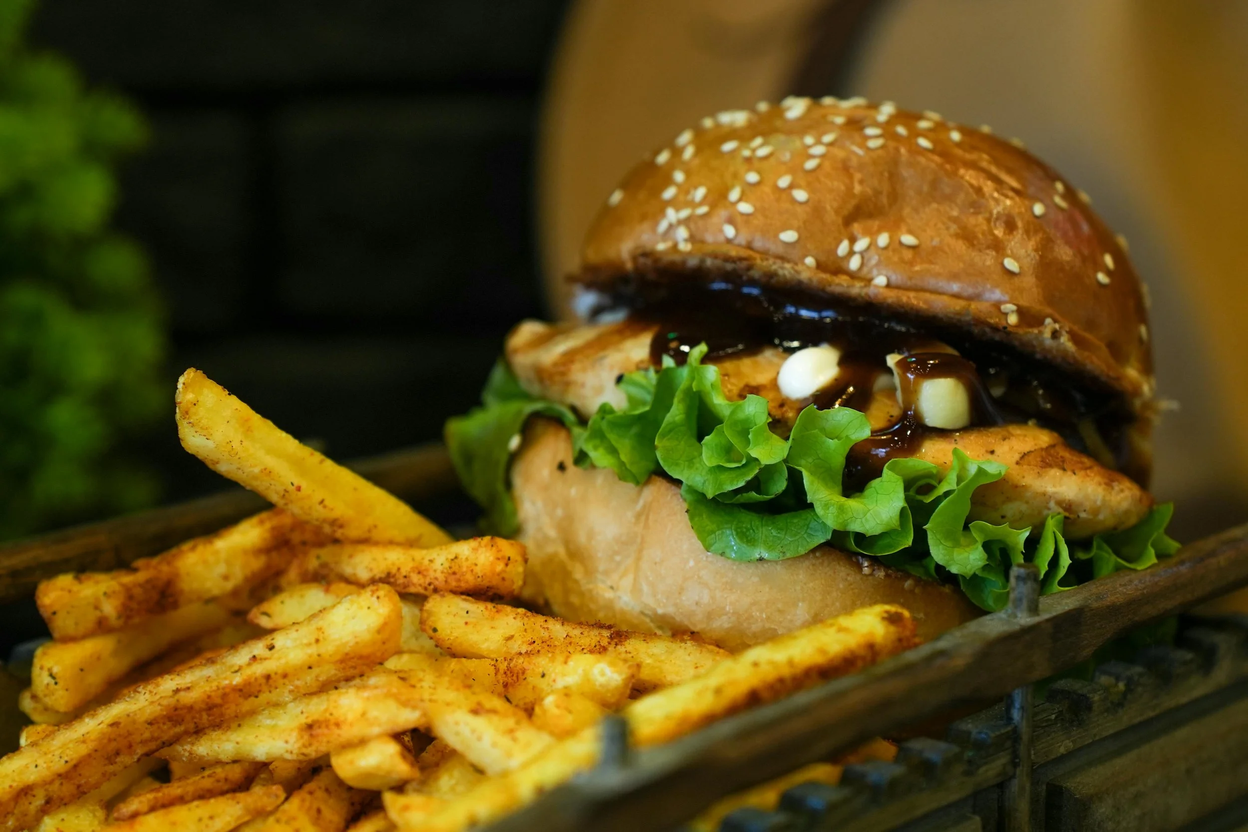 Close-up of a burger with lettuce, grilled chicken, dark sauce, and a sesame seed bun, served with French fries.