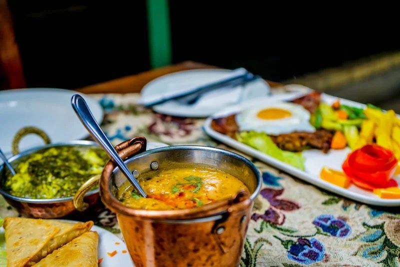 Indian meal setup with curry, rice, roti, and a breakfast plate with eggs and vegetables on a decorative tablecloth.