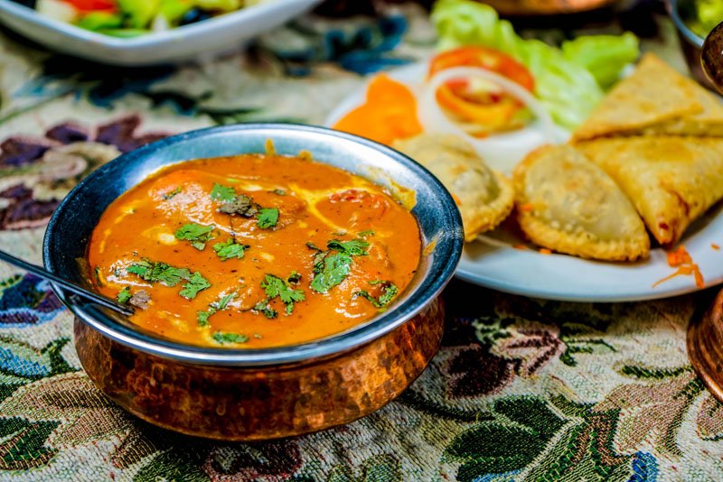 A bowl of Indian curry garnished with cilantro, placed next to a plate with fried snacks and salad on a decorative tablecloth.