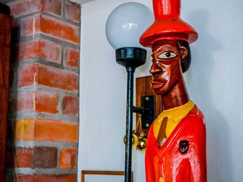 Colorful wooden sculpture of a man with exaggerated features wearing a red jacket and a red hat, standing next to a black floor lamp with a white globe shade.