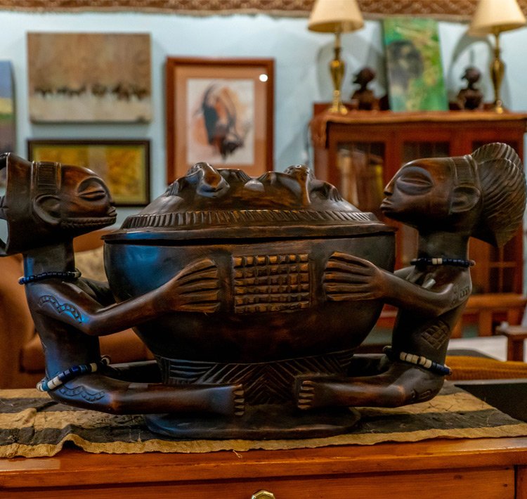 Dark wooden African tribal sculpture of two human figures holding a decorated bowl, sitting on a wooden surface in a room decorated with various paintings and lamps.