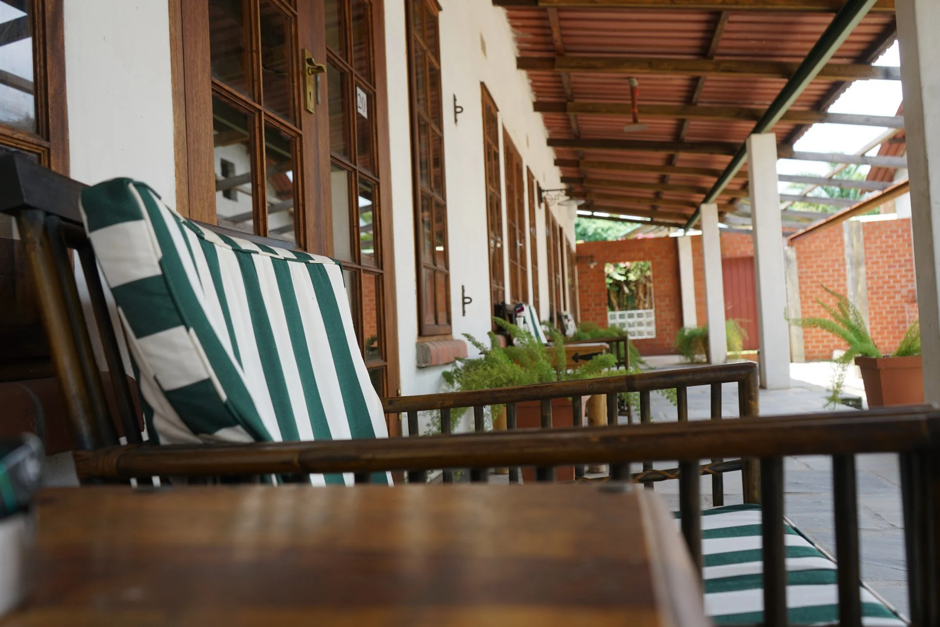 Outdoor patio with wooden chairs featuring green and white striped cushions, potted plants, large windows, and a brick wall in the background.
