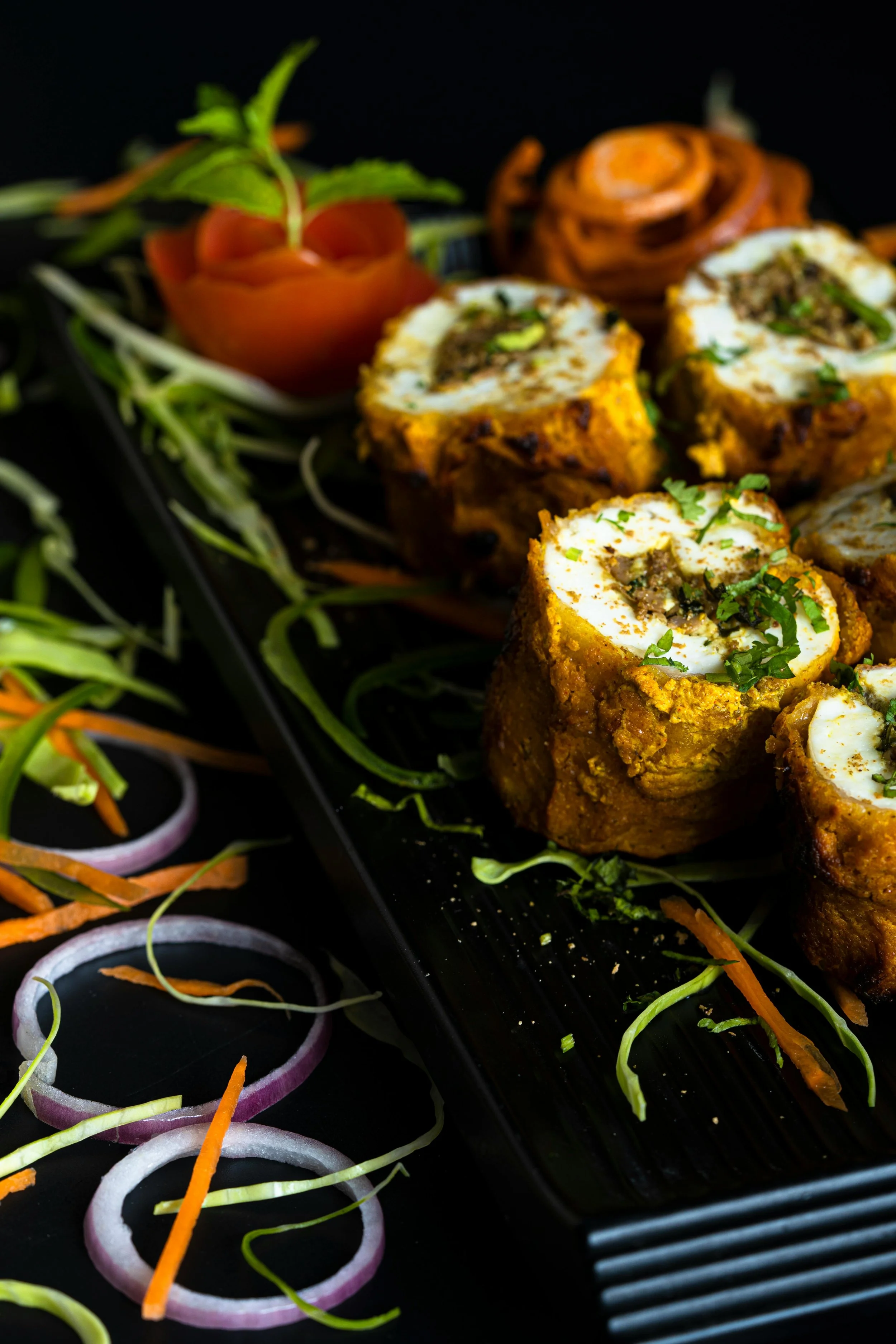 Close-up of stuffed chicken pieces garnished with herbs, with sliced onions, carrots, and a vegetable rose in the background on a black serving platter.
