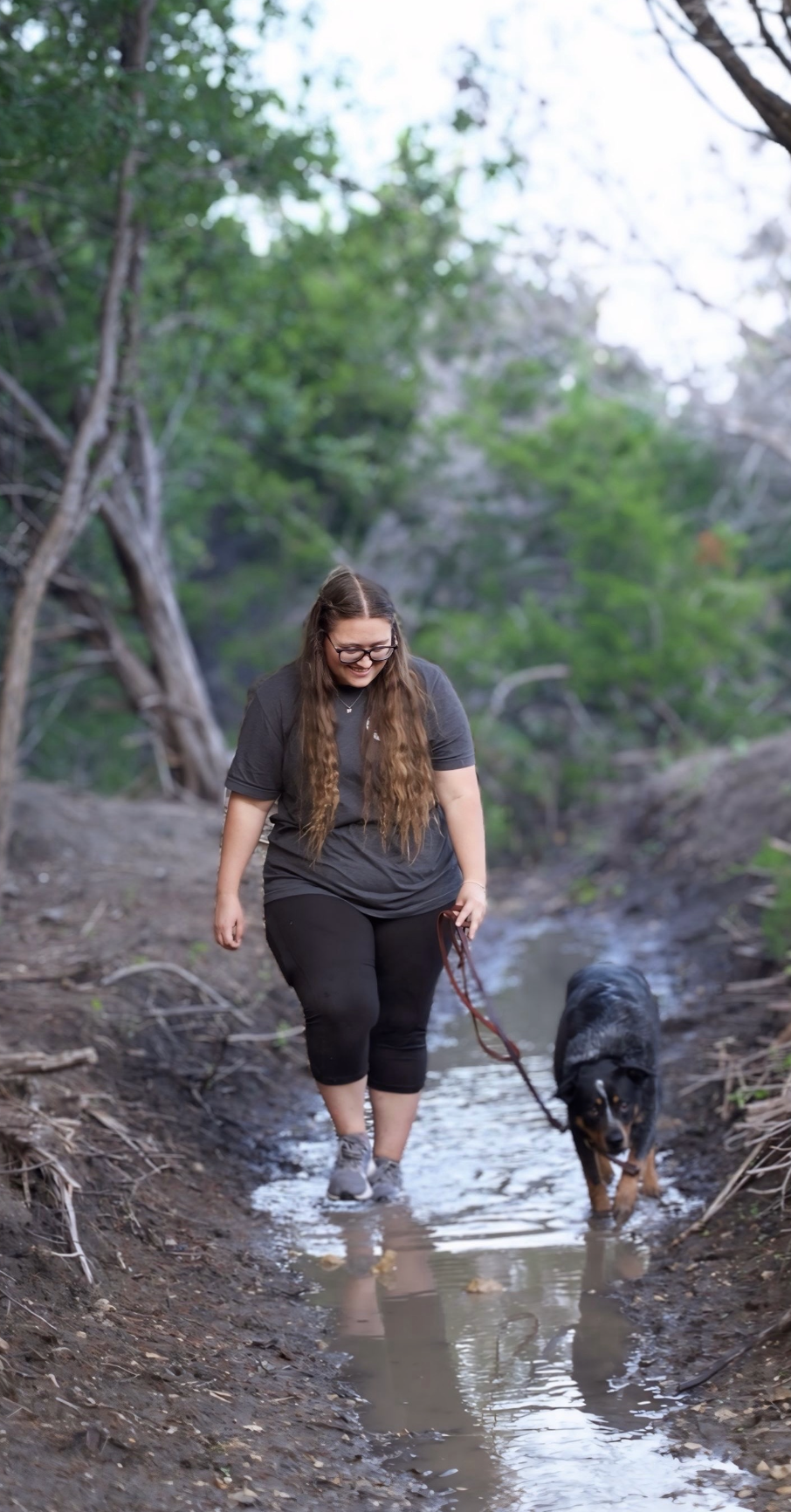 A woman walking through a shallow stream with her dog in a wooded area.