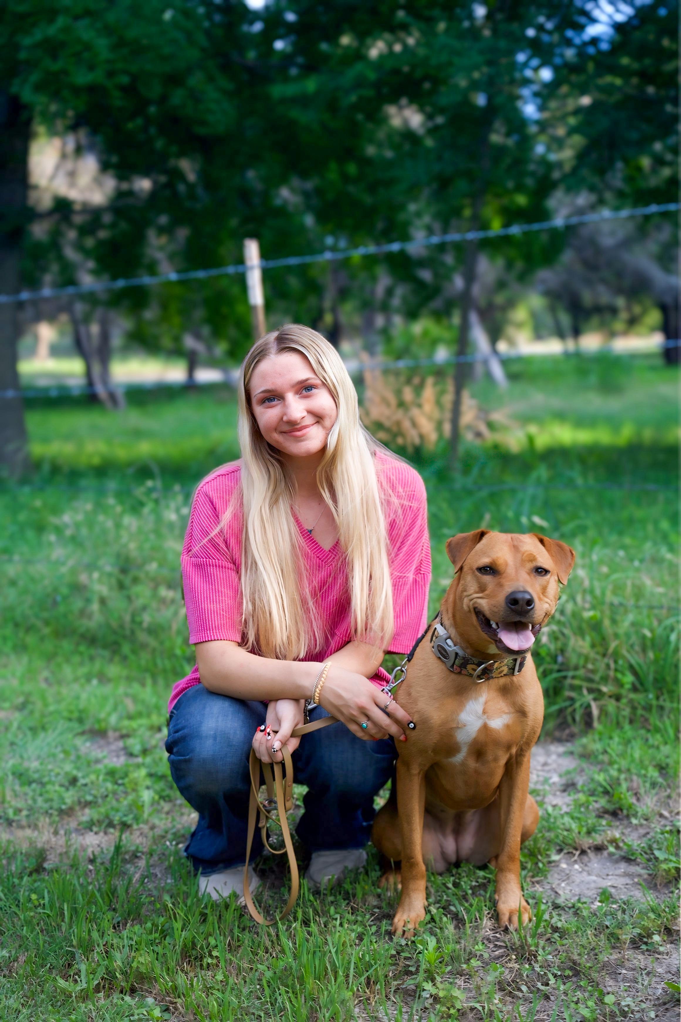 A young woman with long blonde hair wearing a pink top and blue jeans, squatting outdoors on green grass, smiling at the camera with her hand on a brown dog with a black collar, sitting beside her, in a park setting with trees and a fence in the background.