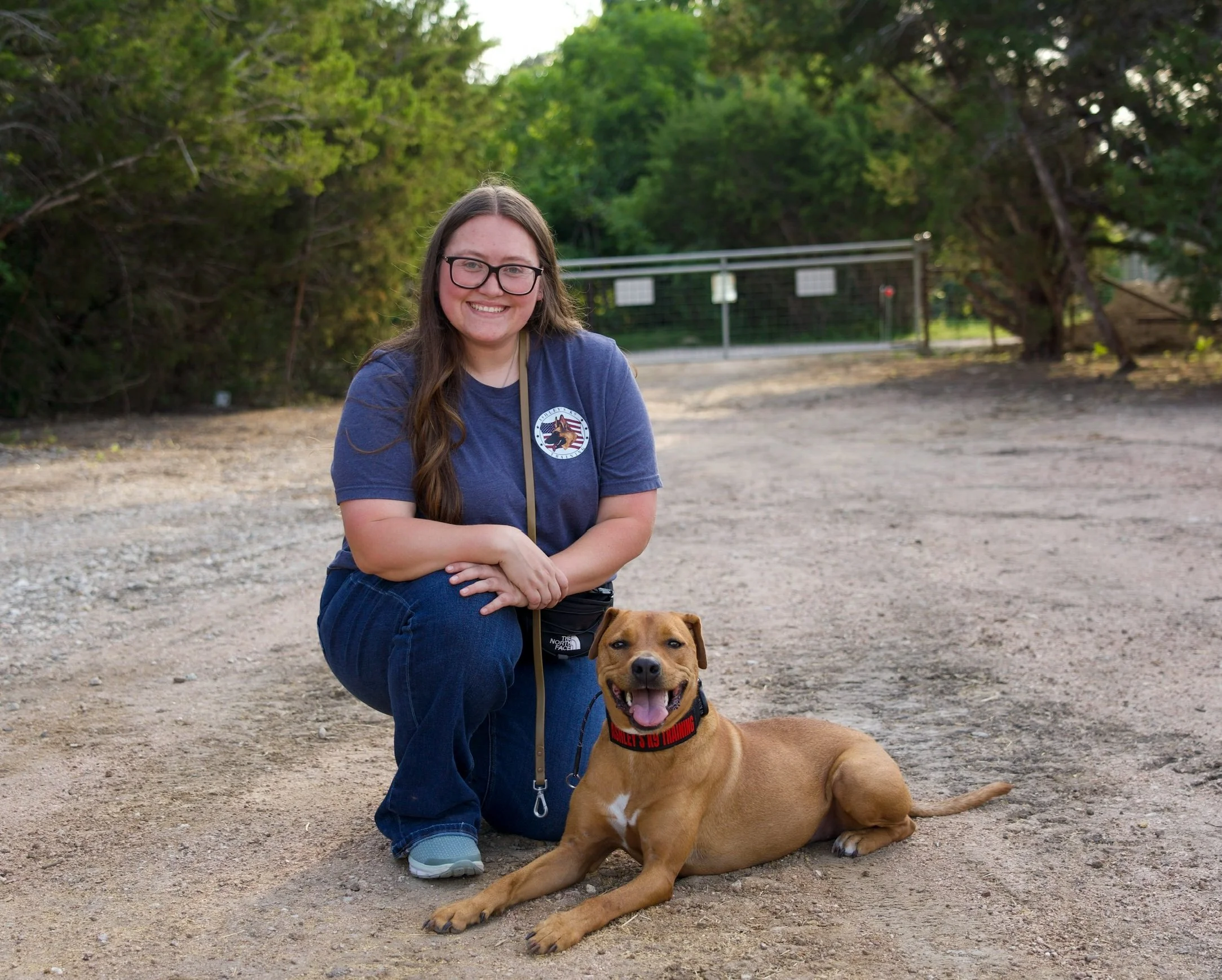 A woman with long hair, glasses, and a blue t-shirt kneeling next to a tan dog with a black harness outside. The woman is smiling, and the dog is sitting with its tongue out. There are green trees and a metal gate in the background.