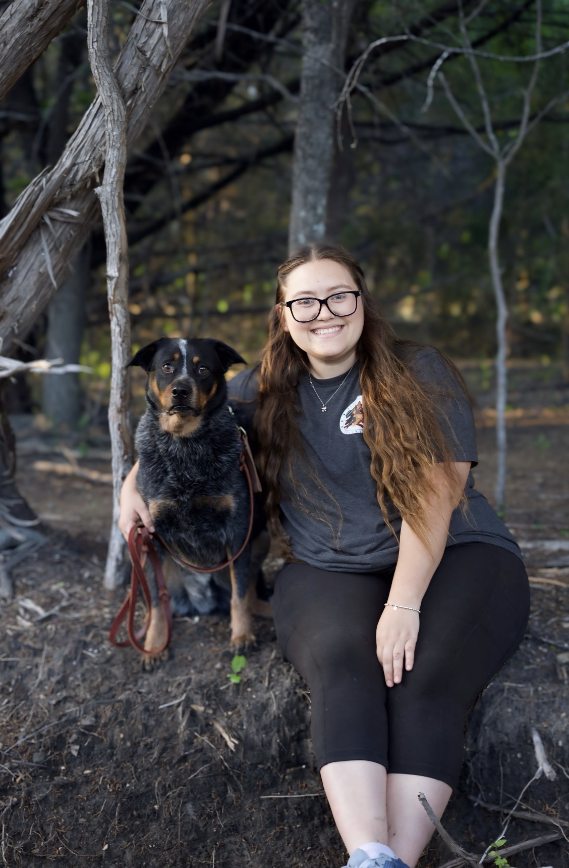 A young woman with long hair and glasses smiling and kneeling next to a black and tan dog in a forest setting.