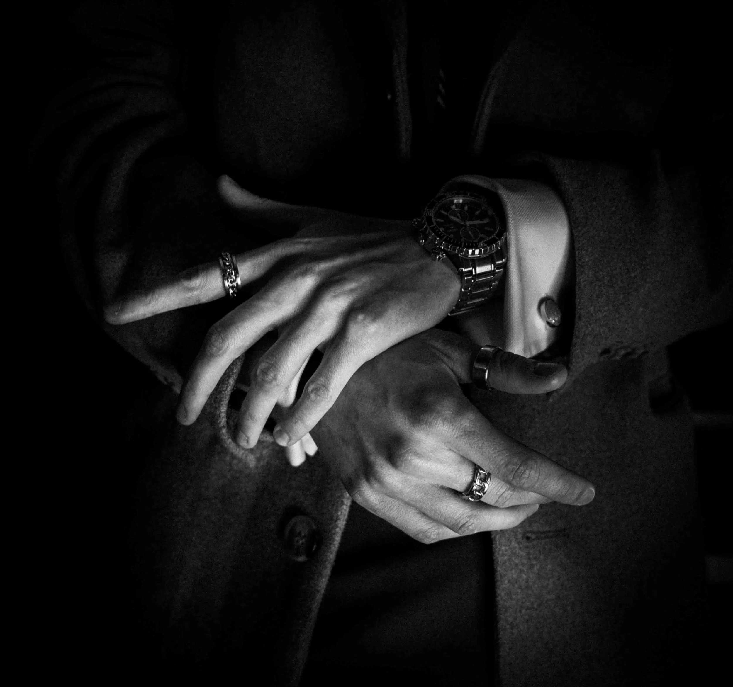 Black and white close-up of two hands with rings and a watch, gently intertwined, suggesting intimacy or connection, against a dark background.