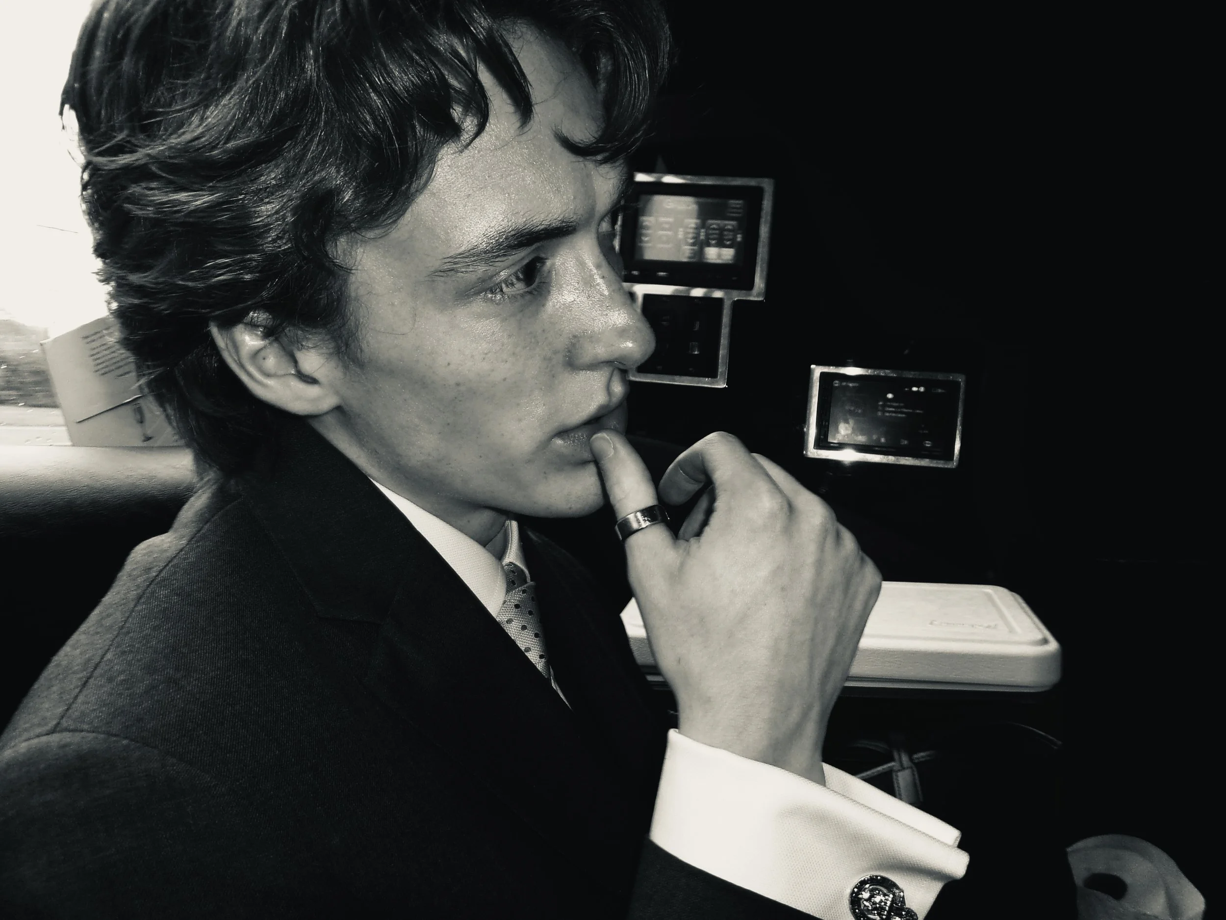 A young man with dark hair and light skin, wearing a suit, looking thoughtful with his hand on his chin, inside a vehicle with electronic devices in the background.