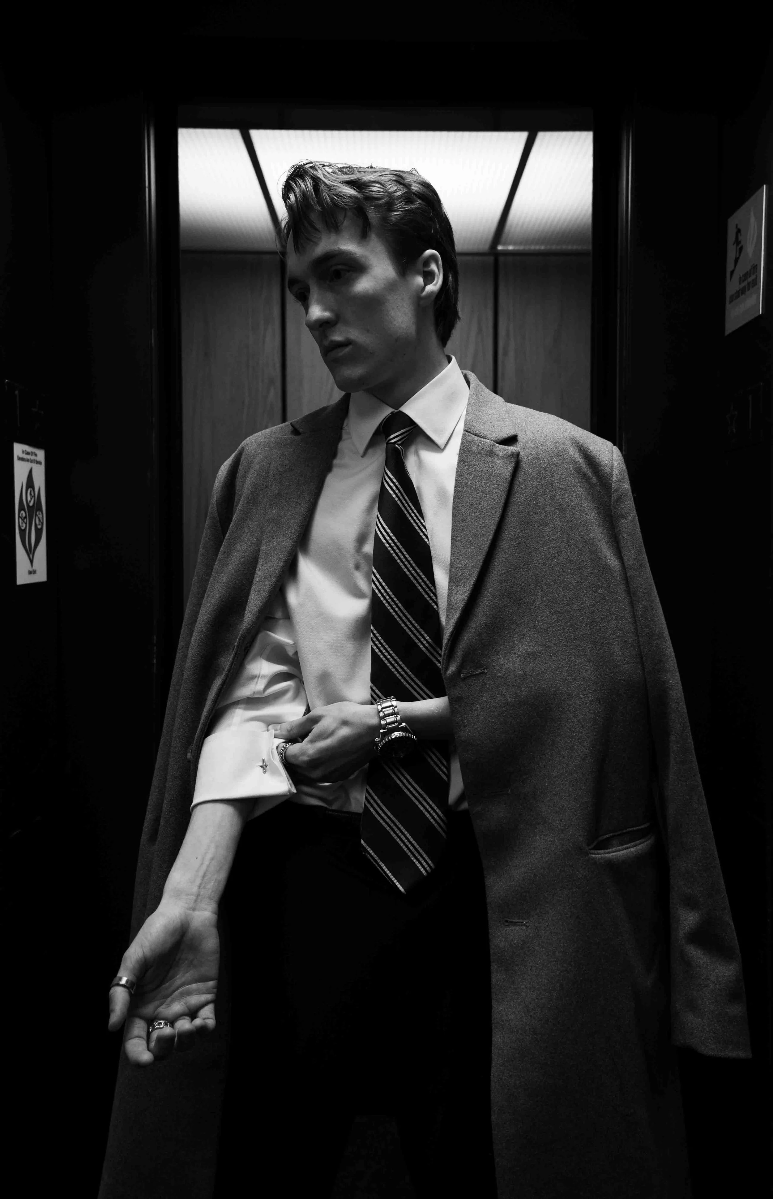 A black-and-white photo of a young man in a suit and tie, with a coat draped over his shoulders, standing inside an elevator with a serious expression.